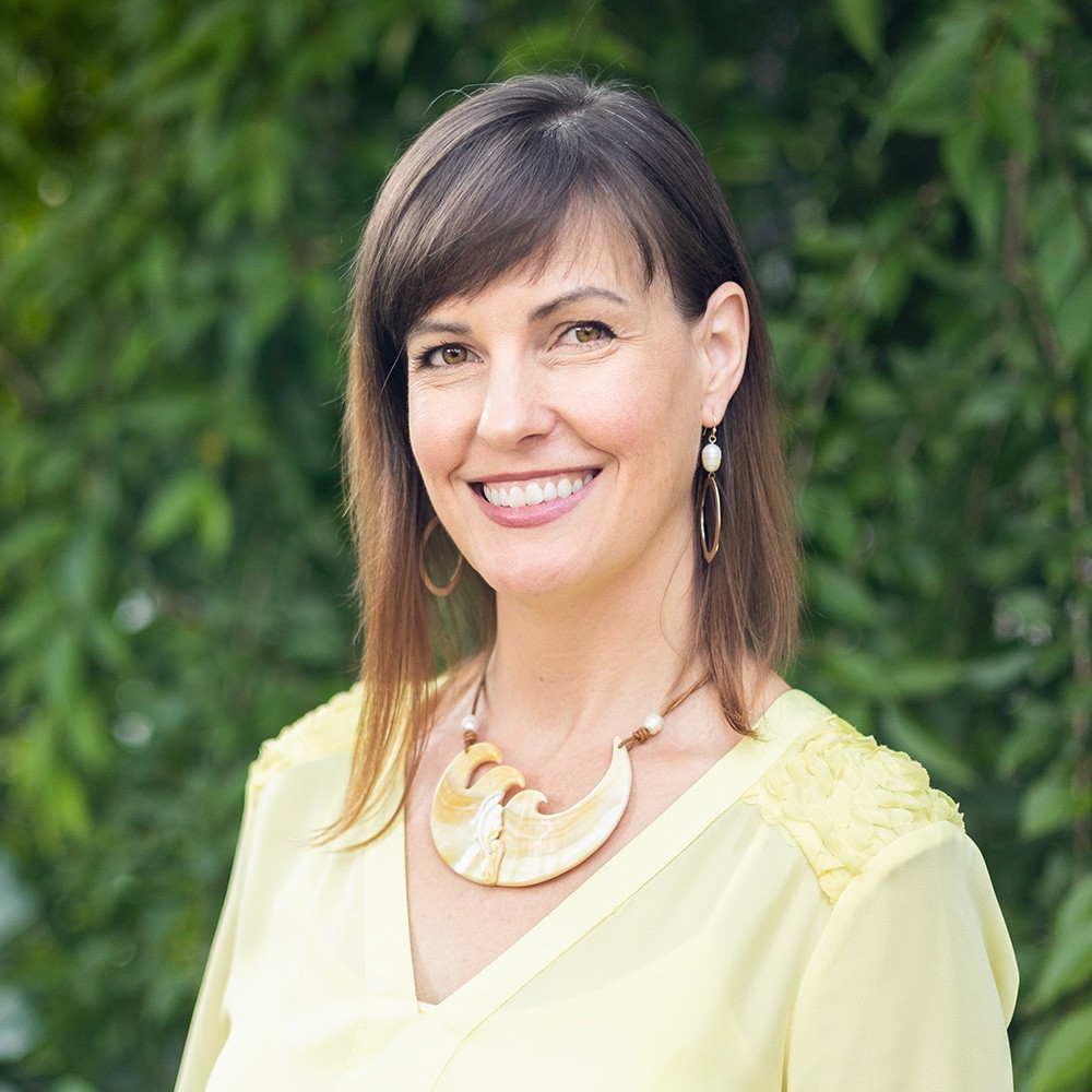 Portrait of Ashley Oros smiling, in a pale yellow blouse against green leaves