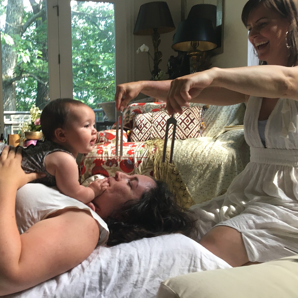 A mother lies back smiling with her baby on her chest while tuning forks are held above them during a session