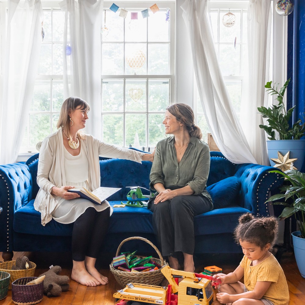 Ashley Oros with a parent on a blue velvet sofa in a sunlit room while a young child plays with toys on the floor